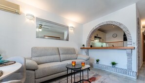 A living room with a sofa, a round coffee table, and a small side table. There is a mirror on the wall, a clock, and a fireplace with a brick arch. - Poolside  2 bedroom apartment sunny retreat   REF052 (Benalmádena)