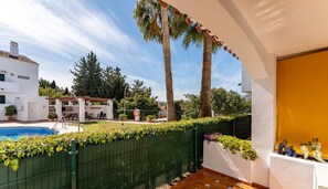 A poolside area with a green fence, a wooden deck, and a view of a building with a white facade and a red roof. - Poolside  2 bedroom apartment sunny retreat   REF052 (Benalmádena)