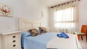 Een slaapkamer met een bed, nachtkastje, stoel en raam met gordijnen.