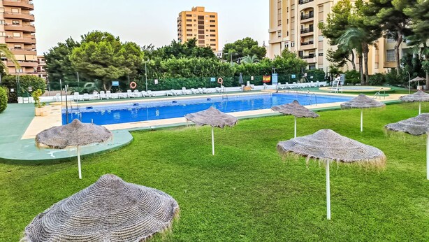 Piscina rodeada por grama e guarda-sóis de palha, com edifícios altos ao fundo.