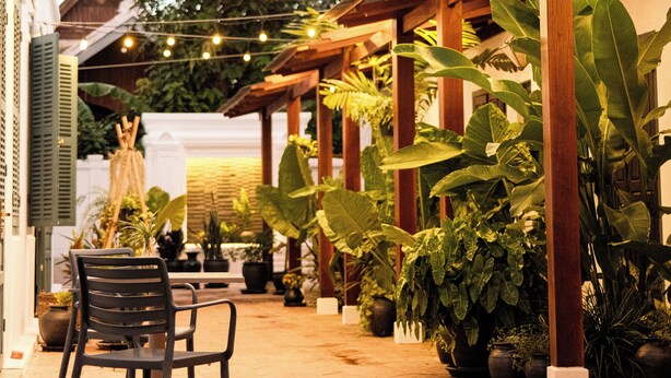 A covered outdoor seating area with wooden pillars, potted plants, and string lights. - Pha Nya Residence (Luang Prabang)