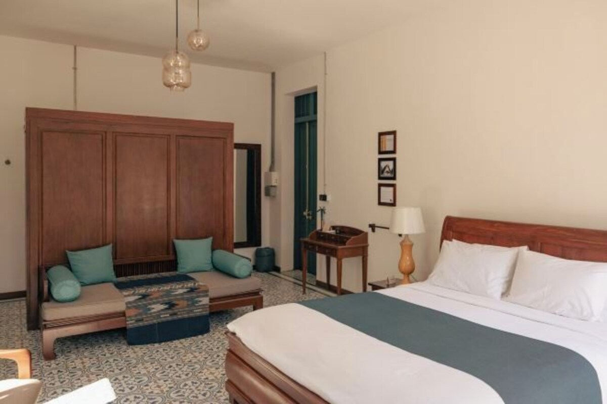 A hotel room with a bed, a sofa with cushions, a wooden headboard, a small table with a lamp, and a door.