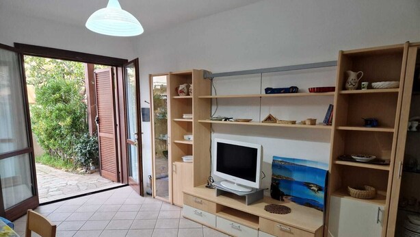 A room with a TV on a wooden stand, a dining table with chairs, and a bookshelf with various items. - Maison Maria, 7 beds (Villasimius)