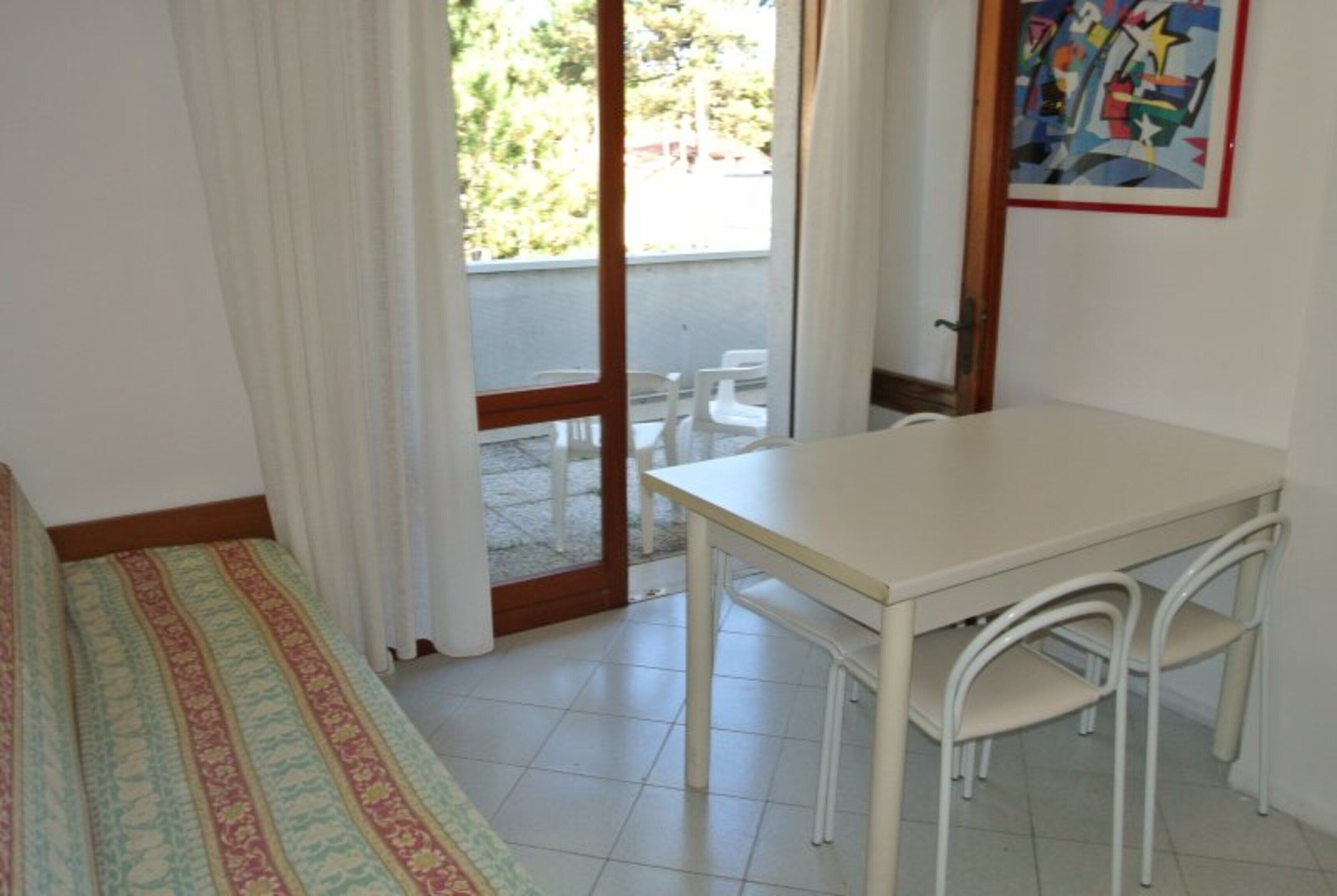 Una habitación con cama, una mesa, dos sillas, una puerta que da a un balcón y un cuadro en la pared.
