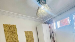 A room with a bed, a white chair, a window with curtains, and wall decorations. - Coquet Appartement À Casablanca (Casablanca)