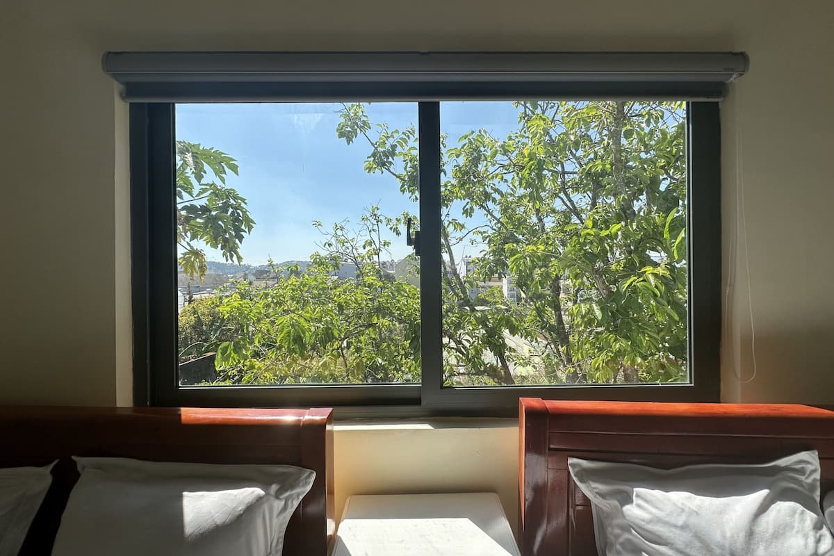 A room with two beds, a window, and a view of trees and buildings.