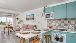 Una cocina moderna con armarios color turquesa, una encimera blanca y un horno empotrado. Hay una zona de comedor con una mesa blanca y taburetes de madera.