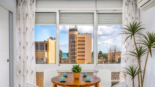 Un comedor moderno con una mesa de madera y sillas blancas, ventanas amplias con cortinas blancas y vistas a un edificio alto.