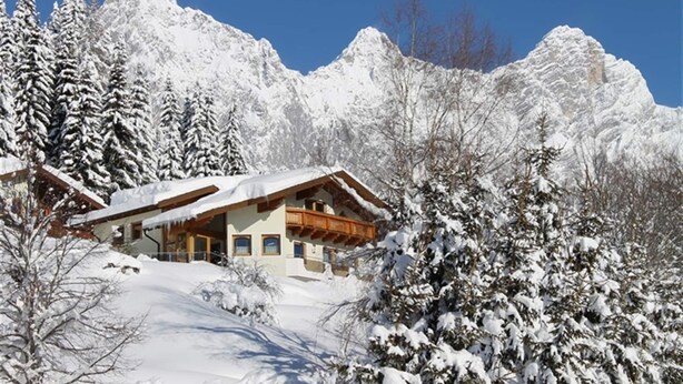 Un chalet de montagne enneigé, avec une façade en bois et de nombreuses fenêtres, entouré d’arbres et de montagnes couverts de neige.
