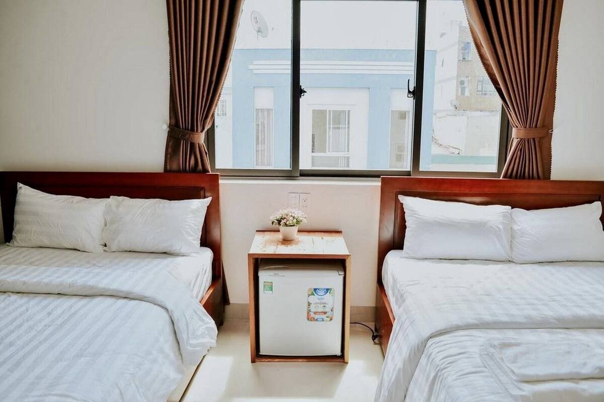 Quarto de hotel com duas camas, uma mesa pequena com um vaso de flores e uma janela com cortinas.