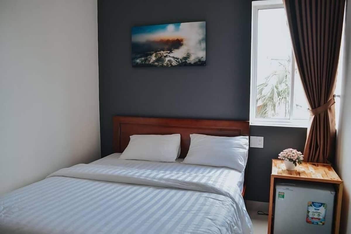 Cama bem arrumada com roupa de cama branca, uma pequena mesa de cabeceira de madeira com um vaso de flores, uma geladeira e um quadro de um vulcão na parede.