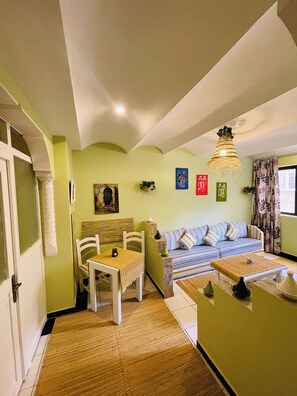 A room with a dining table, chairs, a sofa, a hanging light fixture, and a window with curtains.