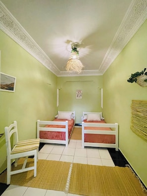 A room with two single beds, a chair, a hanging light fixture, and a wall-mounted decoration.