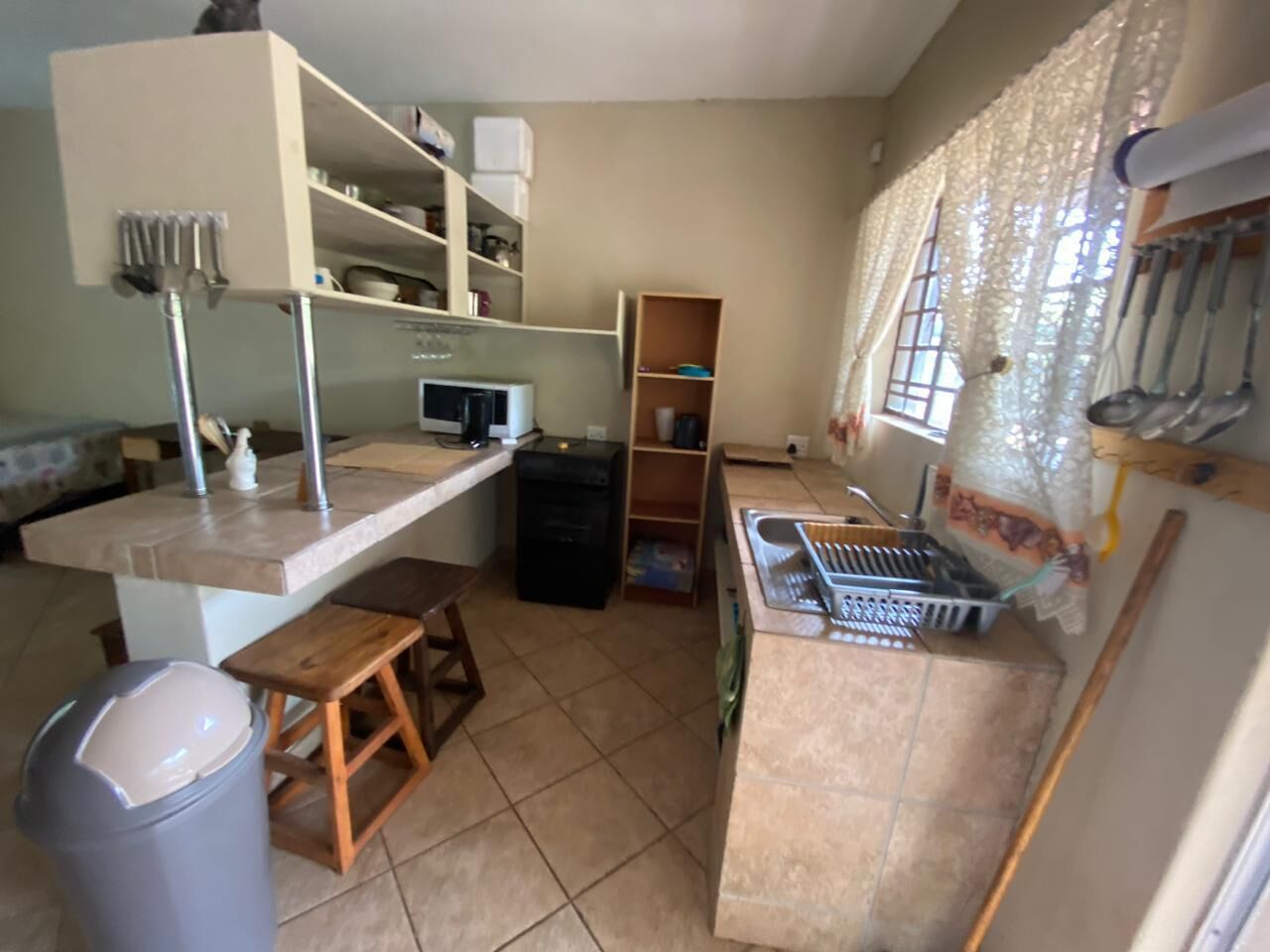 A compact kitchen with a countertop, sink, and a small wooden stool. There is a trash bin and a shelf with kitchen utensils.