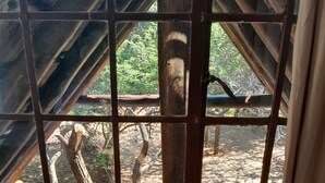Quarto com vista pela janela, com barras de madeira, uma viga de madeira e uma árvore lá fora.