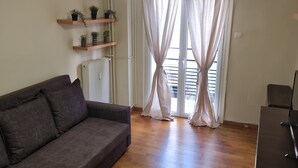 Sala de estar com sofá, piso de madeira, luminária no teto, radiador e janela com cortinas leves.