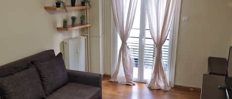 A living room with a sofa, wooden flooring, a ceiling light, a radiator, and a window with sheer curtains.