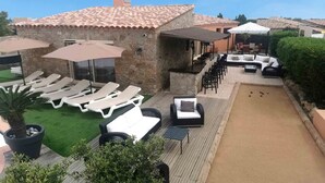 A courtyard with lounge chairs, a bocce court, and a stone building with a tiled roof. - Charmante Maison Avec Piscine et Pétanque (Lumio)
