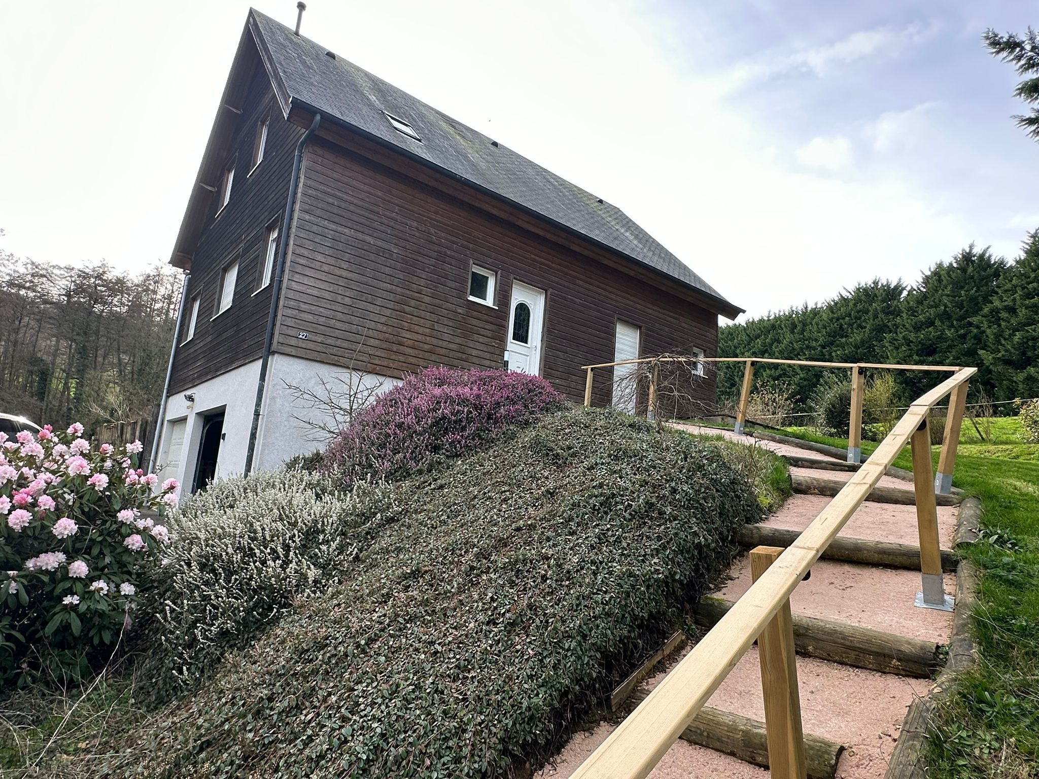 A two-story wooden house with a sloped roof, a wooden staircase with a handrail, and a garden with various shrubs and flowers.