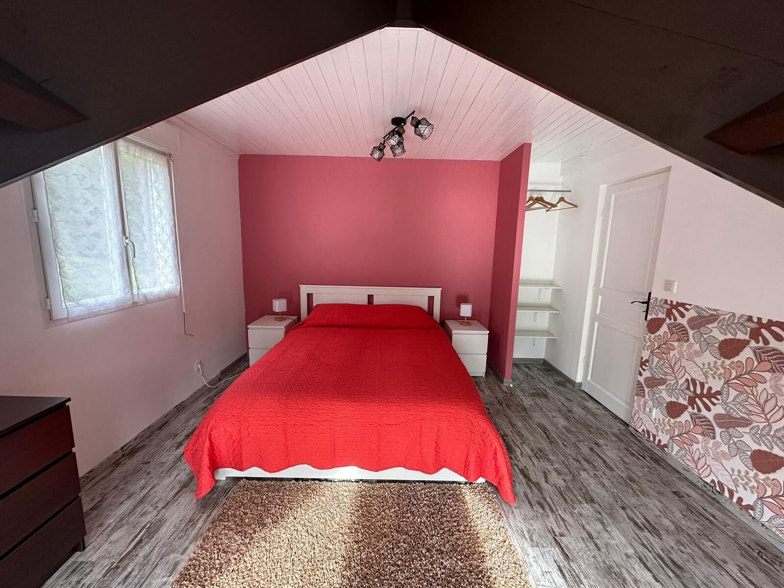 Een slaapkamer met een schuine plafond, een rood beddegoed, een houten dressoir, een behang met patroon en een klein nachtkastje met een lamp.