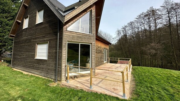 Ein modernes, zweistöckiges Haus mit geneigtem Dach, großen Glasfenstern und einer Holzterrasse.