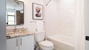 Un baño moderno con ducha de azulejos blancos, encimera de mármol, inodoro y una obra enmarcada sobre el espejo.