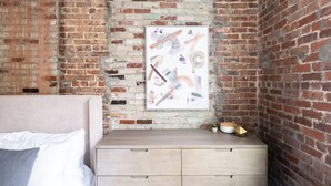 Un dormitorio con pared de ladrillo, una cama con ropa de cama blanca, una mesita de noche de madera con cajones y un cuadro abstracto enmarcado.