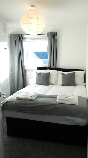 A neatly made bed with a white and gray duvet, grey pillows, and a white throw blanket. There is a window with grey curtains, a white wall, and a hanging light fixture.