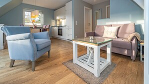 A modern living room with a blue armchair, a glass-top coffee table, a pink sofa, and a kitchen in the background. - Hier Lautet das Motto: Wohnen und Entspannen mit Heimat-atmosphäre (Westerland)