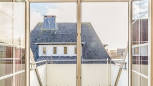 Uitzicht vanaf een balkon, met een heldere lucht, een huis met donker dak en een witte muur met een raam.