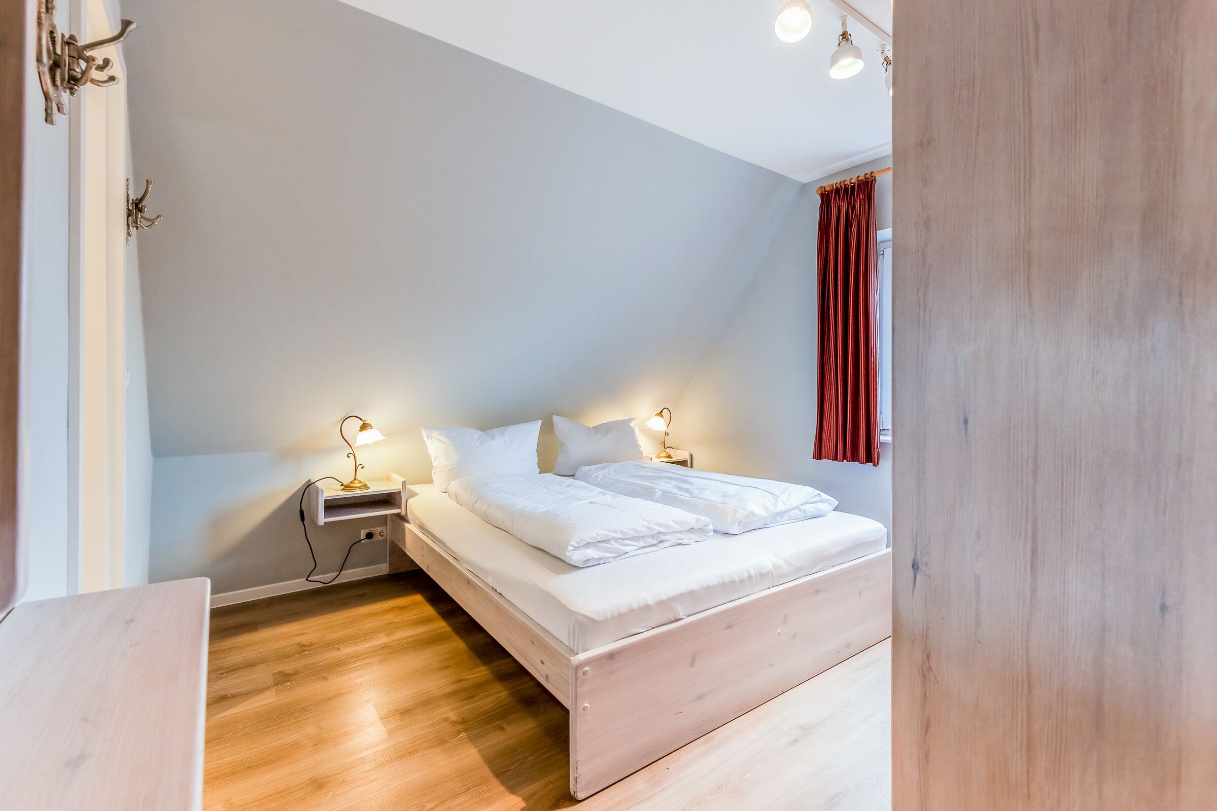 A bedroom with a wooden floor, a bed with white bedding, two bedside lamps, and a nightstand.