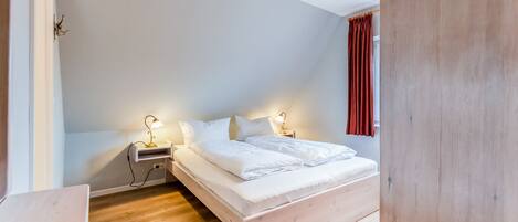 A bedroom with a wooden floor, a bed with white bedding, two bedside lamps, and a nightstand.