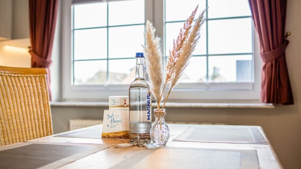 Een tafel met een fles Magnus, een doos Self Homes en een glazen vaas met pampa gras.