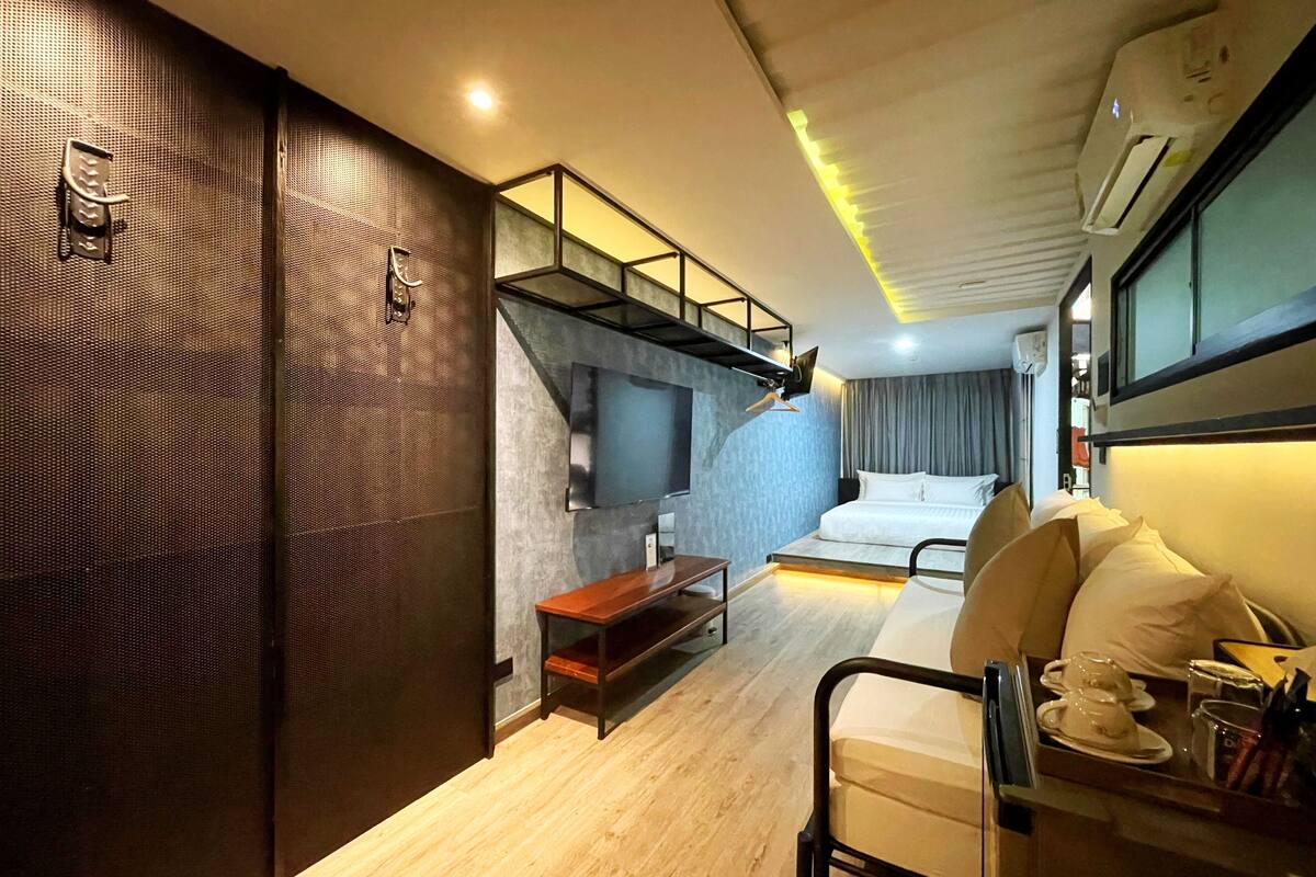 A compact hotel room with a bed, a TV mounted on the wall, a small wooden table, and a seating area with a chair.