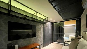 A modern hotel room with a wooden bench, a large TV mounted on the wall, and a large window with a view of the city. - Nights in a Box (Chonburi)