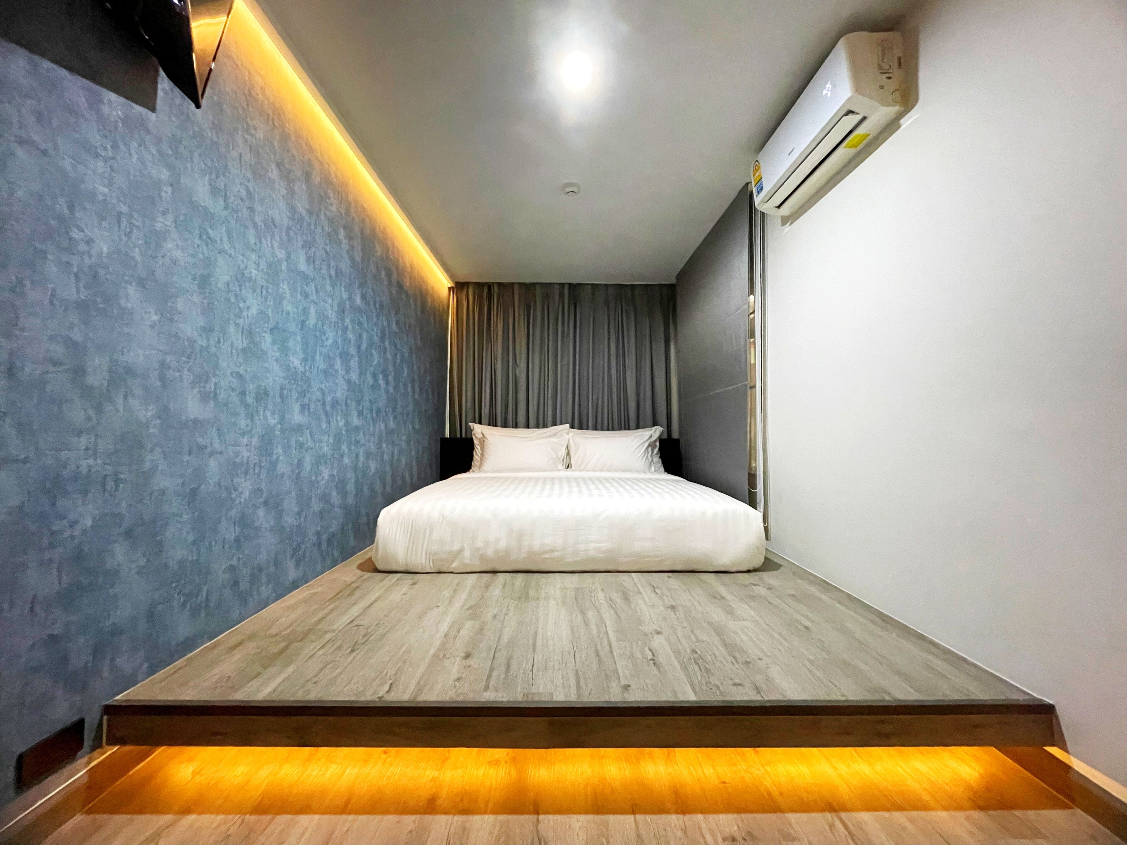 Un dormitorio moderno con una cama centrada sobre una plataforma de madera, una pared gris con textura y un equipo de aire acondicionado en la pared.