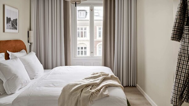 Ett hotellrum med en säng, en hängande lampa, ett fönster med gardiner och ett rutigt plagg som hänger på väggen.