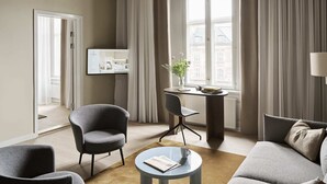 Une chambre d’hôtel moderne dotée d’une grande fenêtre, d’un bureau agrémenté d’un vase de fleurs, de deux fauteuils, d’un canapé et d’un téléviseur fixé au mur.
