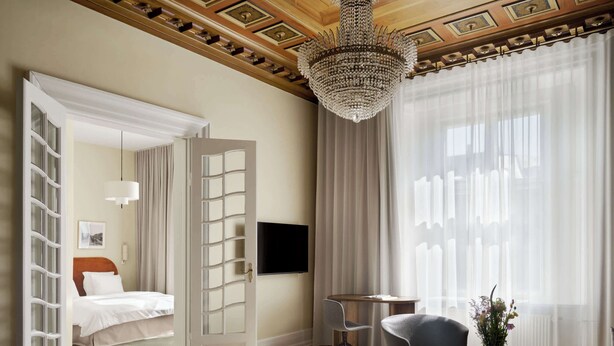 Une chambre d’hôtel spacieuse, dotée d’un grand lit, d’une télévision, d’un lustre et offrant une vue sur la ville à travers de fines rideaux.
