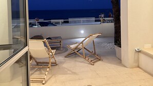 Ein Balkon mit Blick auf das Meer im Abendlicht, Holzmöbel und eine Palme.