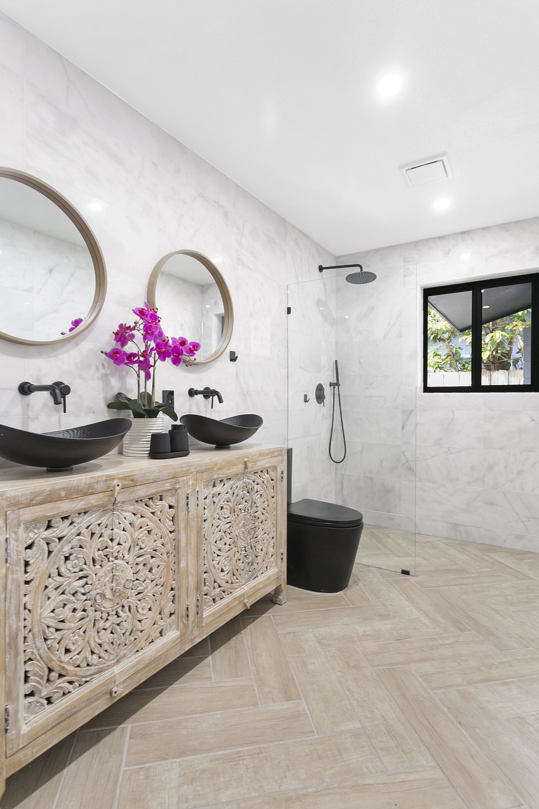 A modern bathroom with two large oval sinks, a freestanding toilet, a shower, and a window with a view of greenery.