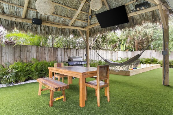 An outdoor patio with a wooden table and chairs, a hammock, and a grill.