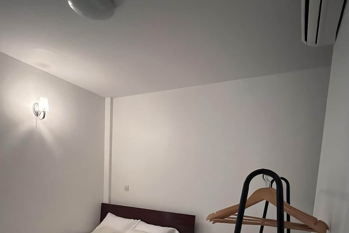 Una cama bien tendida con sábanas blancas, una mesita de noche de madera con dos cajones, una lámpara fijada a la pared y un perchero de madera sobre un soporte.
