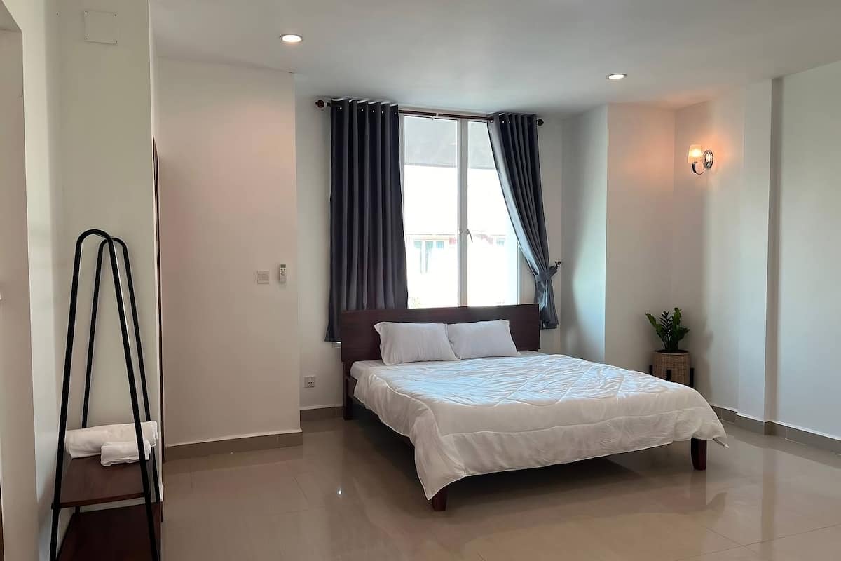 Una habitación de hotel moderna con una cama grande, una mesita de noche pequeña, una planta en maceta y una ventana con cortinas.