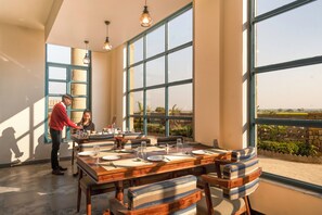 A dining area with a large table set for four, large windows, and a view of the outdoors. - Jay Gurudev Palpur Fort Kuno By Paandora Collections (Karahal)