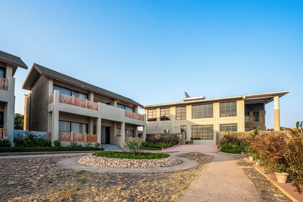 A modern building with multiple floors, a central courtyard with a stone pathway, and surrounding greenery. - Jay Gurudev Palpur Fort Kuno By Paandora Collections (Karahal)