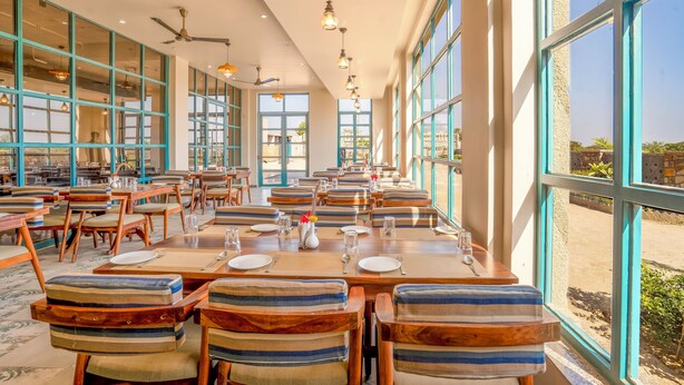 A restaurant with long tables, striped cushioned chairs, and large windows offering a view of the outdoors. - Jay Gurudev Palpur Fort Kuno By Paandora Collections (Karahal)
