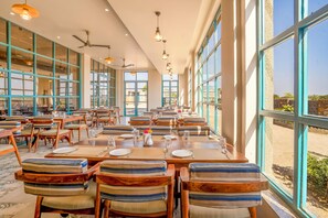 A restaurant with long tables, striped cushioned chairs, and large windows offering a view of the outdoors. - Jay Gurudev Palpur Fort Kuno By Paandora Collections (Karahal)