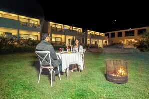 A dining area with a table and chairs, a fire pit, and a building with multiple balconies in the background. - Jay Gurudev Palpur Fort Kuno By Paandora Collections (Karahal)
