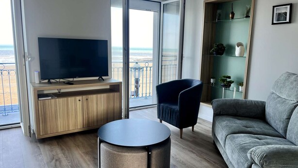 Un saló modern amb televisió, una taula de centre rodona, un sofà i una cadira. Hi ha un balcó amb vista al mar.